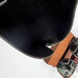 May include: A black, curved chair seat with a damaged edge, resting on a wooden stand. Tortoiseshell sunglasses are placed on the stand. The chair seat has a textured surface.