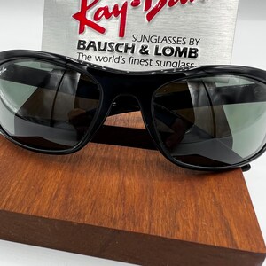 May include: Black Ray-Ban sunglasses with dark lenses. The sunglasses are displayed on a wooden surface, with a silver sign in the background that reads "Ray-Ban" and "Sunglasses by Bausch & Lomb".