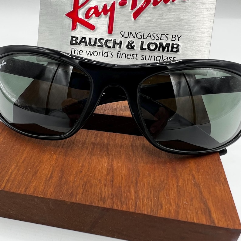 May include: Black Ray-Ban sunglasses with dark lenses. The sunglasses are displayed on a wooden surface, with a silver sign in the background that reads "Ray-Ban" and "Sunglasses by Bausch & Lomb".