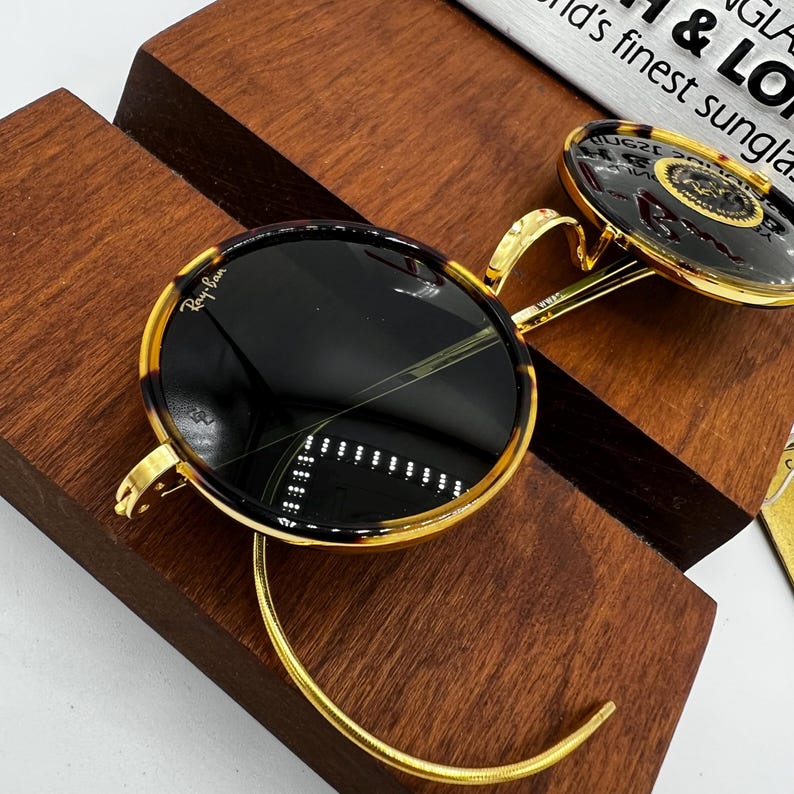 K&ouml;nnte beinhalten: Runde Sonnenbrille mit Schildpattmuster-Rahmen und goldfarbenen Metallakzenten. Die Gl&auml;ser sind dunkel, und die Sonnenbrille liegt auf einem Holzdisplay. Der Markenname "Ray-Ban" ist sichtbar.