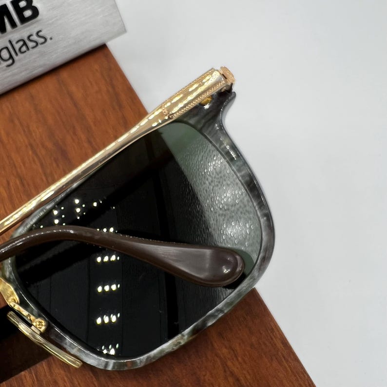 Pu&ograve; includere: Occhiali da sole con montatura marmorizzata grigia e nera, aste color oro e lenti scure. Gli occhiali da sole sono appoggiati su una superficie di legno. Il testo "MB glass" &egrave; visibile sullo sfondo.