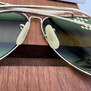 K&ouml;nnte beinhalten: Goldene Aviator-Sonnenbrille mit gr&uuml;nen Gl&auml;sern und wei&szlig;en Nasenpads. Die Sonnenbrille liegt auf einer Holzoberfl&auml;che.