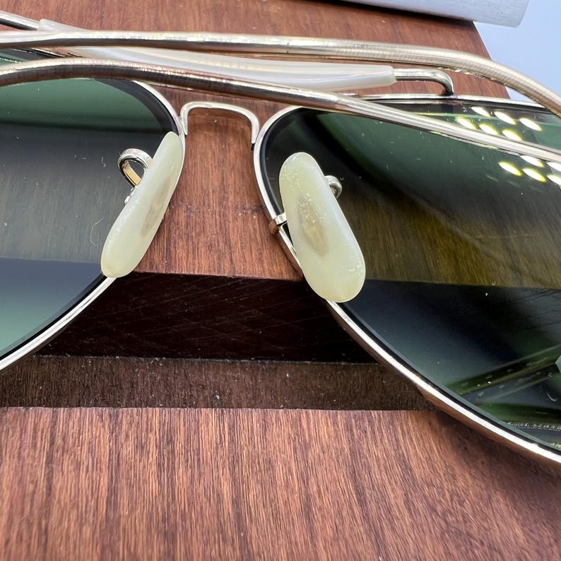 K&ouml;nnte beinhalten: Goldene Aviator-Sonnenbrille mit gr&uuml;nen Gl&auml;sern und wei&szlig;en Nasenpads. Die Sonnenbrille liegt auf einer Holzoberfl&auml;che.