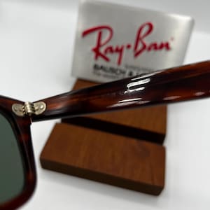 Puede incluir: Primer plano de gafas de sol Ray-Ban con montura de carey y lentes verdes. El logotipo de Ray-Ban es visible en la patilla. Las gafas de sol descansan sobre un expositor de madera, con el logotipo de Ray-Ban en el fondo.