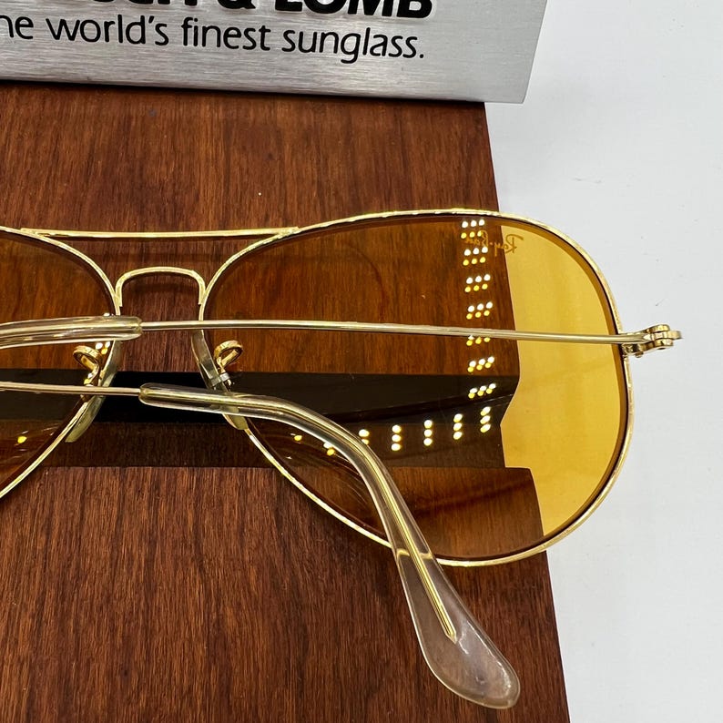 Puede incluir: Gafas de sol tipo aviador en tono dorado con lentes tintadas en &aacute;mbar y monturas de metal fino. La lente derecha muestra el logotipo de Ray-Ban en blanco. Un letrero plateado arriba dice Bausch & Lomb y the world's finest sunglass.