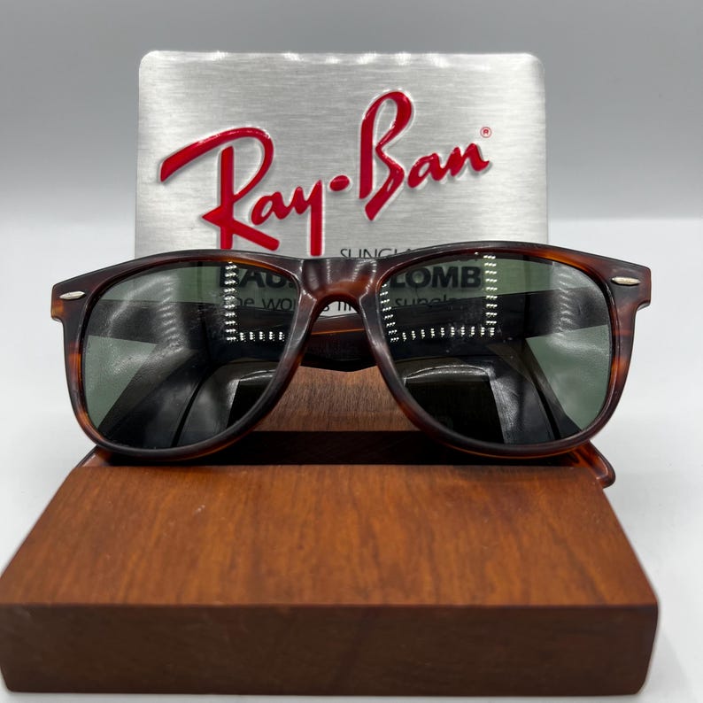 May include: Tortoiseshell-framed sunglasses with green lenses are displayed on a wooden stand. The Ray-Ban logo is visible on a silver background behind the sunglasses. The sunglasses have a classic, rectangular shape.