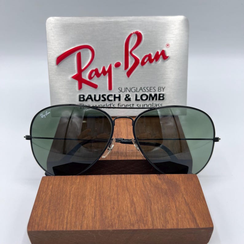 Pu&ograve; includere: Occhiali da sole aviator Ray-Ban neri con lenti verdi. Gli occhiali sono esposti su un supporto in legno con un cartello in metallo argentato che recita "Ray-Ban Sunglasses by Bausch & Lomb, the world's finest sunglass".