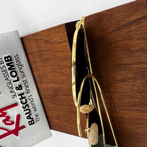 Pode incluir: &Oacute;culos de sol com arma&ccedil;&atilde;o dourada e lentes pretas. Os &oacute;culos de sol t&ecirc;m um design &uacute;nico com uma arma&ccedil;&atilde;o dourada curva e detalhes decorativos. O texto na caixa prateada diz "BAUSCH & LOMB Sunglasses By The world's finest sunglass."