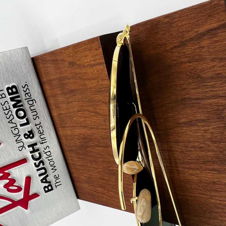Pode incluir: &Oacute;culos de sol com arma&ccedil;&atilde;o dourada e lentes pretas. Os &oacute;culos de sol t&ecirc;m um design &uacute;nico com uma arma&ccedil;&atilde;o dourada curva e detalhes decorativos. O texto na caixa prateada diz "BAUSCH & LOMB Sunglasses By The world's finest sunglass."