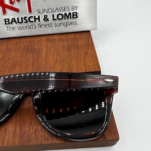 Puede incluir: Gafas de sol marrones oscuras con un acabado brillante. Las gafas de sol se exhiben sobre una superficie de madera. El texto "SUNGLASSES BY BAUSCH & LOMB The world's finest sunglass" es visible en el fondo.