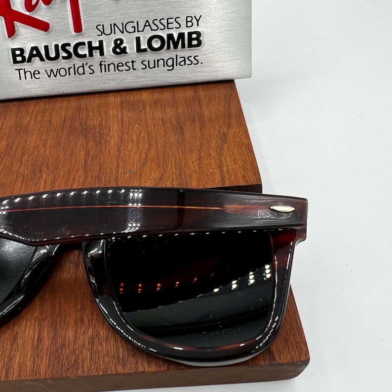 Puede incluir: Gafas de sol marrones oscuras con un acabado brillante. Las gafas de sol se exhiben sobre una superficie de madera. El texto "SUNGLASSES BY BAUSCH & LOMB The world's finest sunglass" es visible en el fondo.