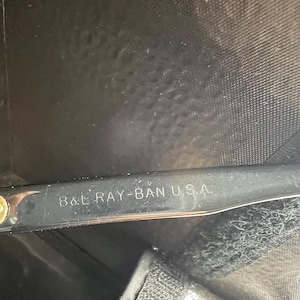 May include: Close-up of a black eyeglass case with the text "8 & L RAY-BAN U.S.A." printed in white. The case has a textured surface and a velcro closure.
