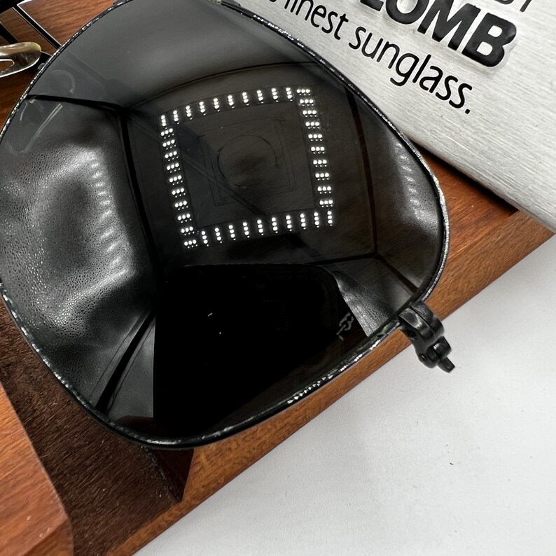 K&ouml;nnte beinhalten: Schwarze Sonnenbrille mit einem quadratischen Muster aus kleinen wei&szlig;en Lichtern in den Gl&auml;sern. Die Sonnenbrille liegt auf einer Holzoberfl&auml;che, mit dem Text "finest sunglass" im Hintergrund.