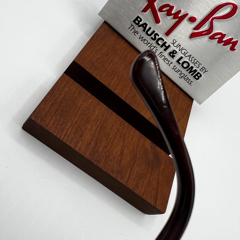 May include: A close-up of a wooden display stand with a dark brown stripe and a silver sign that reads "Ray-Ban SUNGLASSES BY BAUSCH & LOMB The world's finest sunglasses." The arm of a pair of sunglasses is visible.