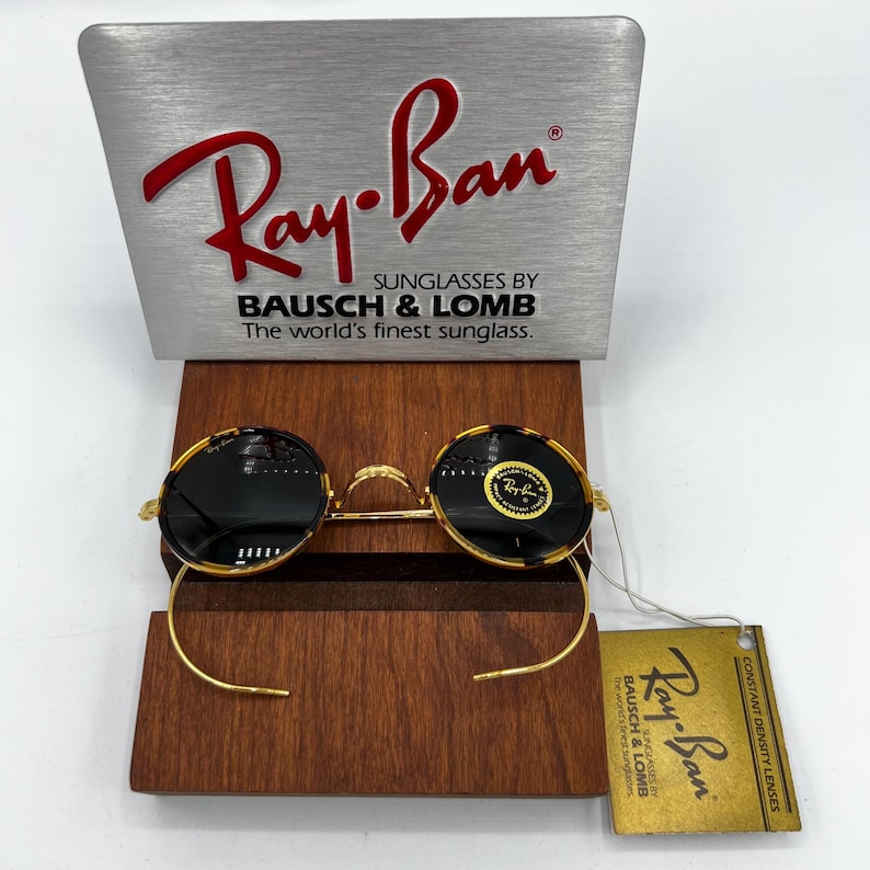 K&ouml;nnte beinhalten: Runde Ray-Ban Sonnenbrille mit goldenem Rahmen und dunklen Gl&auml;sern, pr&auml;sentiert auf einem Holzst&auml;nder. Das Ray-Ban-Logo ist auf den Gl&auml;sern und dem St&auml;nder sichtbar. Die Sonnenbrille hat ein Etikett mit dem Ray-Ban-Logo und Text.
