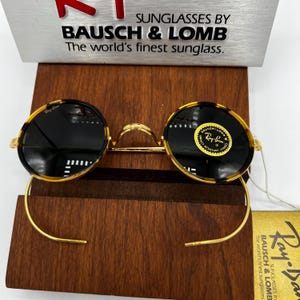 K&ouml;nnte beinhalten: Runde Sonnenbrille mit einem Rahmen im Schildpattmuster und dunklen Gl&auml;sern. Der goldfarbene Metallrahmen hat einen Steg und gebogene B&uuml;gel. Die Sonnenbrille wird auf einem Holzst&auml;nder pr&auml;sentiert. Der Text "SUNGLASSES BY BAUSCH & LOMB" ist sichtbar.