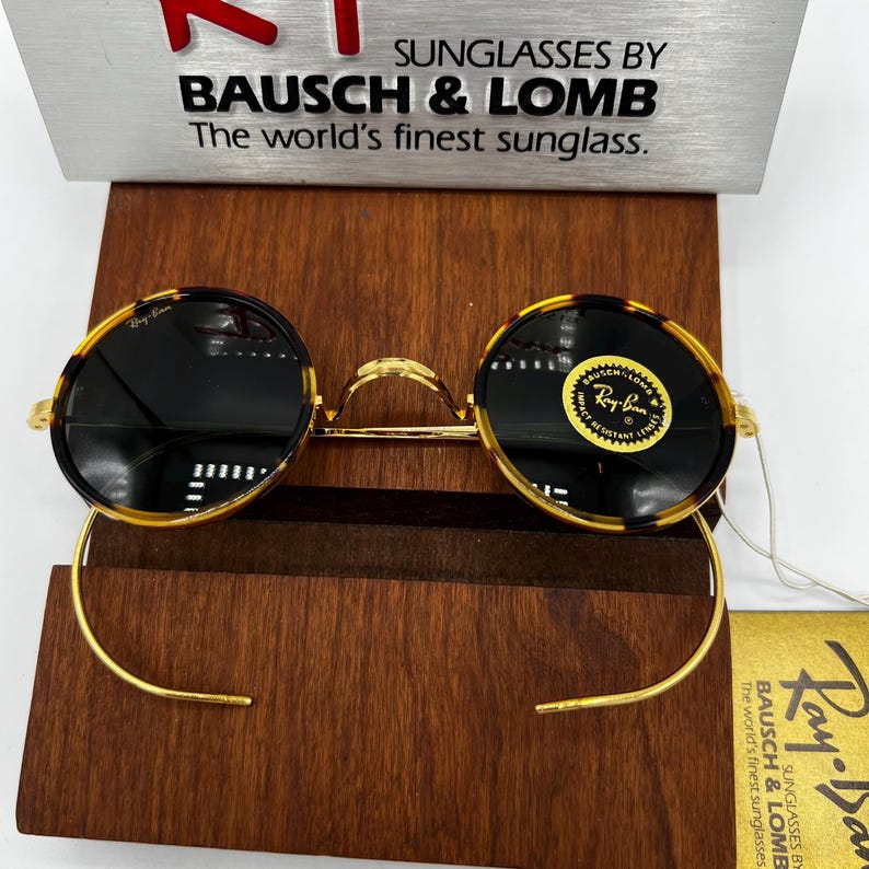 K&ouml;nnte beinhalten: Runde Sonnenbrille mit einem Rahmen im Schildpattmuster und dunklen Gl&auml;sern. Der goldfarbene Metallrahmen hat einen Steg und gebogene B&uuml;gel. Die Sonnenbrille wird auf einem Holzst&auml;nder pr&auml;sentiert. Der Text "SUNGLASSES BY BAUSCH & LOMB" ist sichtbar.