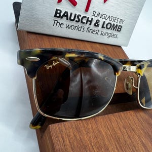 May include: Ray-Ban sunglasses with a tortoiseshell pattern on the upper frame and temples. The lower frame is gold-colored. The sunglasses are displayed on a wooden surface, with a metal sign that reads "BAUSCH & LOMB" and "The world's finest sunglass."