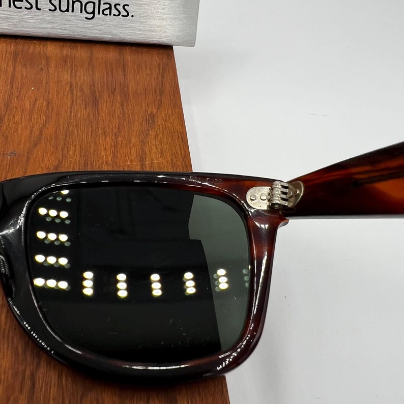 Puede incluir: Primer plano de gafas de sol con montura negra y lentes oscuras. Las patillas tienen un dise&ntilde;o de carey. La bisagra es plateada. Las gafas de sol est&aacute;n sobre una superficie de madera.