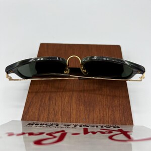 Pu&ograve; includere: Occhiali da sole Ray-Ban con lenti verde scuro e montatura marmorizzata nera e verde. Gli occhiali hanno dettagli in metallo color oro e sono esposti su un supporto in legno. Il logo Ray-Ban &egrave; visibile in rosso.
