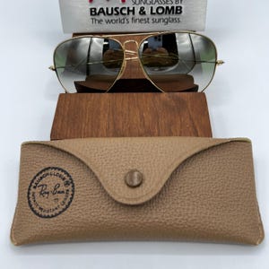 K&ouml;nnte beinhalten: Goldene Aviator-Sonnenbrille mit gr&uuml;nen Gl&auml;sern und einem braunen Lederetui. Das Etui hat ein rundes Logo mit der Aufschrift "Ray-Ban" und "Impact Resistant Lenses".