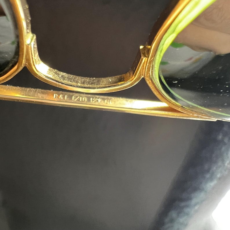 May include: Close-up of gold-rimmed sunglasses with green lenses. The frame has a classic design with a bridge and arms. The text "R&L (210 12x46" is visible on the frame.