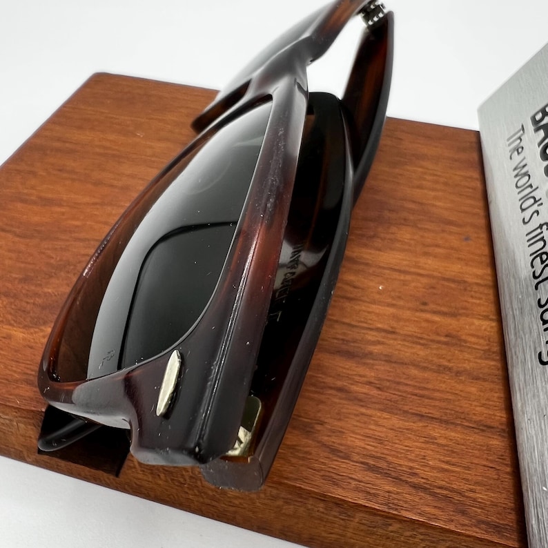 May include: Dark brown sunglasses with a tortoiseshell pattern. The sunglasses have dark lenses and are resting on a wooden display stand. The frame has a rectangular shape.