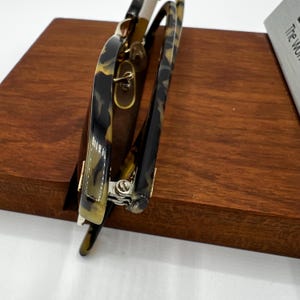 May include: A pair of eyeglasses with a tortoiseshell pattern on the frames. The glasses have a gold-colored hinge and are resting on a wooden surface. The lenses are clear, and the overall design is classic and stylish.