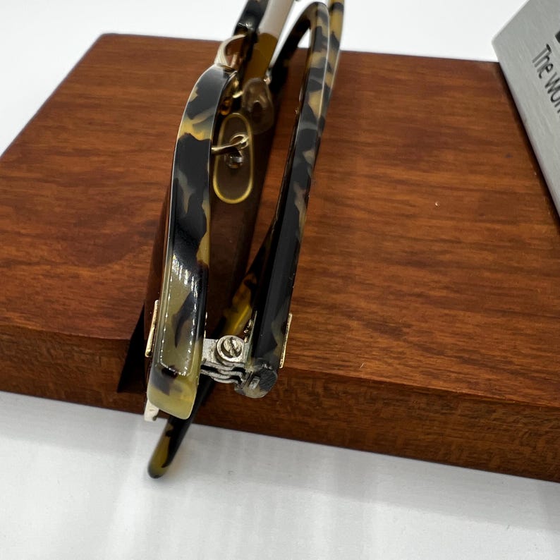 May include: A pair of eyeglasses with a tortoiseshell pattern on the frames. The glasses have a gold-colored hinge and are resting on a wooden surface. The lenses are clear, and the overall design is classic and stylish.