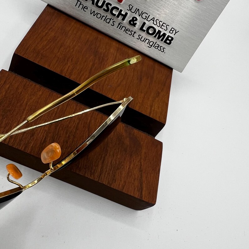 May include: Gold-rimmed sunglasses with orange nose pads are displayed on a wooden stand. The sunglasses are partially visible, with the text "SUNGLASSES BY BAUSCH & LOMB" on a silver sign in the background. The world's finest sunglass.