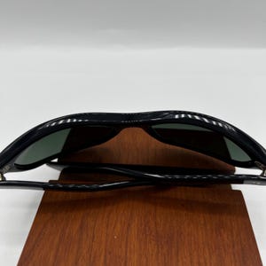 May include: Black sunglasses with dark green lenses. The sunglasses have a classic, rectangular frame design. The image shows the sunglasses resting on a wooden surface, showcasing their sleek and modern aesthetic.