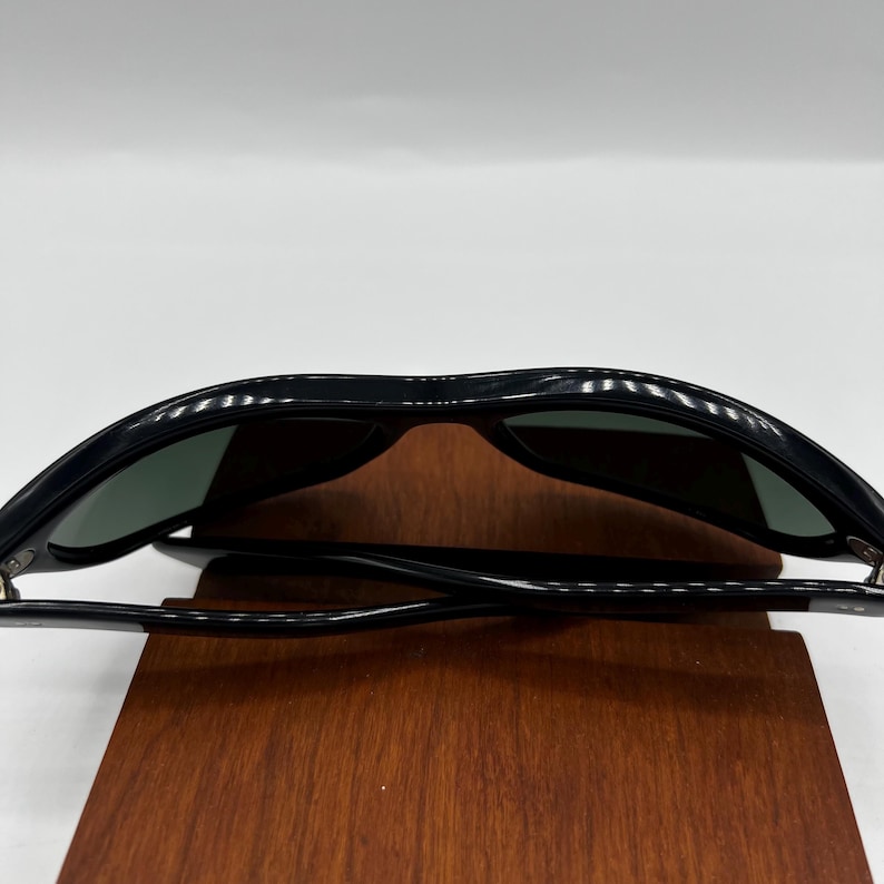 May include: Black sunglasses with dark green lenses. The sunglasses have a classic, rectangular frame design. The image shows the sunglasses resting on a wooden surface, showcasing their sleek and modern aesthetic.