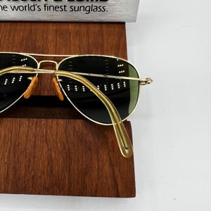 May include: Gold-rimmed sunglasses with green lenses and orange nose pads. The sunglasses are displayed on a dark wooden stand. The text "the world's finest sunglass" is visible at the top of the image.