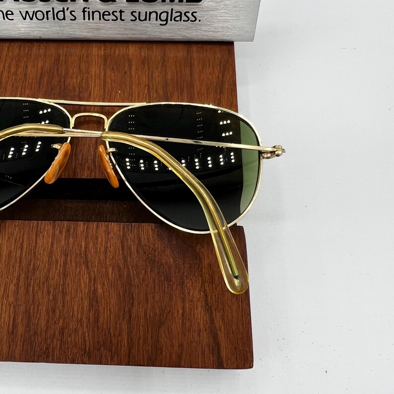 May include: Gold-rimmed sunglasses with green lenses and orange nose pads. The sunglasses are displayed on a dark wooden stand. The text "the world's finest sunglass" is visible at the top of the image.