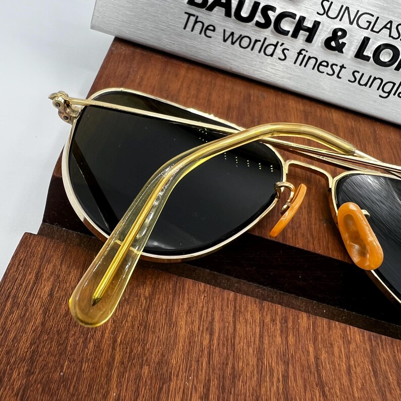 May include: Gold-rimmed sunglasses with dark lenses and orange nose pads. The temples are a transparent yellow. The text "BAUSCH & LOMB The world's finest sunglasses" is visible in the background.