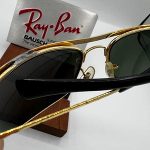 Puede incluir: Gafas de sol con montura dorada, lentes verde oscuro y patillas negras. El logotipo de Ray-Ban es visible en un expositor plateado. Las gafas de sol se exhiben en un soporte de madera.