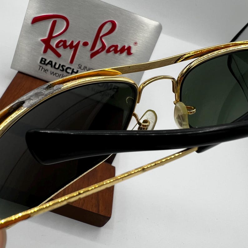 Puede incluir: Gafas de sol con montura dorada, lentes verde oscuro y patillas negras. El logotipo de Ray-Ban es visible en un expositor plateado. Las gafas de sol se exhiben en un soporte de madera.