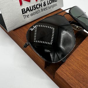 K&ouml;nnte beinhalten: Schwarze Ray-Ban Sonnenbrille mit reflektierender Oberfl&auml;che, auf einem Holzst&auml;nder. Die Sonnenbrille hat ein klassisches Aviator-Design mit silbernem Steg und dunklen Gl&auml;sern. Der Text "BAUSCH & LOMB The world's finest sunglass" ist sichtbar.