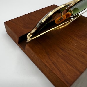 May include: A wooden wine bottle holder with a gold-colored metal frame. The holder is designed to cradle a wine bottle, with a slot in the wooden base. The frame has decorative elements and holds the bottle at an angle.