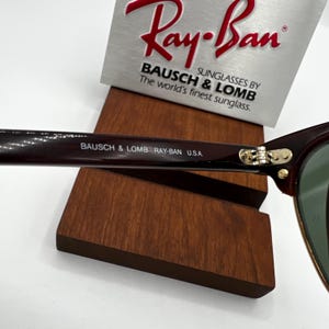 May include: Close-up of Ray-Ban sunglasses with a dark brown frame and green lenses. The text "BAUSCH & LOMB RAY-BAN U.S.A." is printed on the arm. The Ray-Ban logo is visible on a silver display stand.