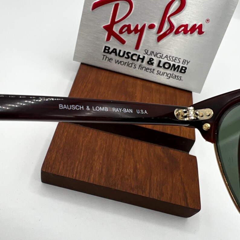May include: Close-up of Ray-Ban sunglasses with a dark brown frame and green lenses. The text "BAUSCH & LOMB RAY-BAN U.S.A." is printed on the arm. The Ray-Ban logo is visible on a silver display stand.