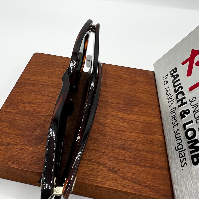 Puede incluir: Gafas de sol carey con un patr&oacute;n marr&oacute;n oscuro y &aacute;mbar, apoyadas sobre una superficie de madera. Las gafas de sol est&aacute;n parcialmente abiertas, revelando la montura interior. El texto "BAUSCH & LOMB The world's finest sunglass." es visible.
