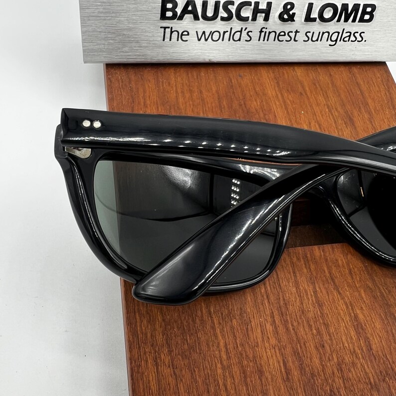 May include: Black sunglasses with dark lenses. The sunglasses are displayed on a wooden surface. The text "BAUSCH & LOMB The world's finest sunglass." is visible above the sunglasses.