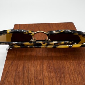 May include: Sunglasses with a tortoise shell pattern frame and gold-colored metal accents. The lenses are a warm brown color. The sunglasses are resting on a wooden surface.