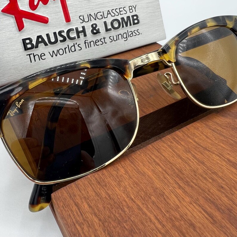 May include: Ray-Ban sunglasses with a tortoiseshell frame and gold accents. The lenses are a warm brown color. The sunglasses are displayed on a wooden surface, with the text "SUNGLASSES BY BAUSCH & LOMB The world's finest sunglass."
