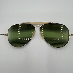 K&ouml;nnte beinhalten: Goldene Aviator-Sonnenbrille mit gr&uuml;nen Gl&auml;sern. Die Sonnenbrille hat einen d&uuml;nnen goldenen Metallrahmen und eine goldene Metallbr&uuml;cke.
