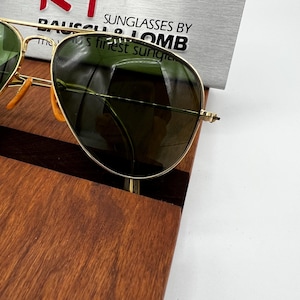 May include: Gold-framed sunglasses with green lenses. The sunglasses are displayed on a wooden surface. The text "SUNGLASSES BY LOMB" is visible in the background, along with the phrase "the finest sunglasses".