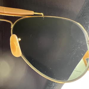 May include: Gold-framed aviator sunglasses with dark lenses. The sunglasses have a classic teardrop shape, with a gold metal frame and brown temple tips. The nose pads are a light tan color. The lenses are dark, providing shade and protection from the sun.