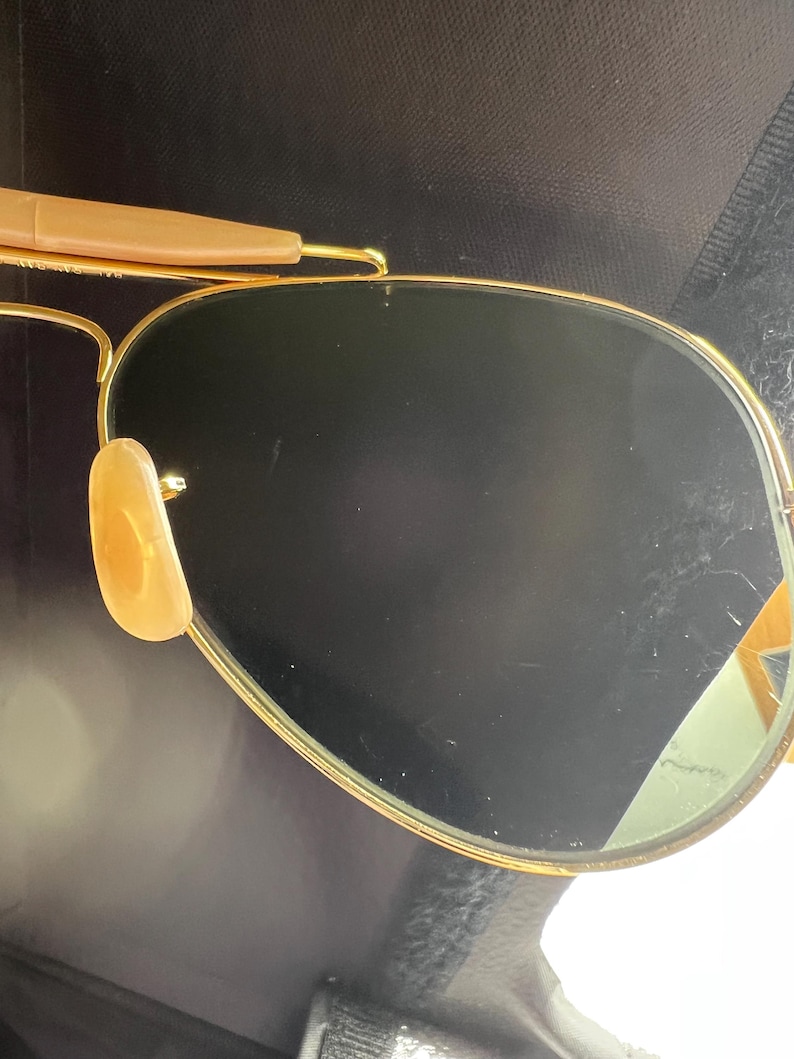 May include: Gold-framed aviator sunglasses with dark lenses. The sunglasses have a classic teardrop shape, with a gold metal frame and brown temple tips. The nose pads are a light tan color. The lenses are dark, providing shade and protection from the sun.