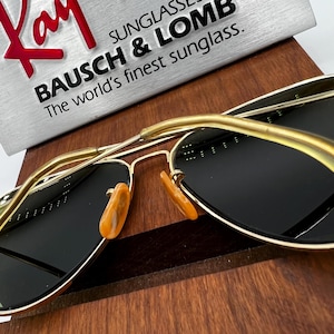 May include: Gold-framed sunglasses with dark lenses and orange nose pads. The sunglasses are displayed on a wooden surface, with the text "BAUSCH & LOMB The world's finest sunglass" visible in the background.